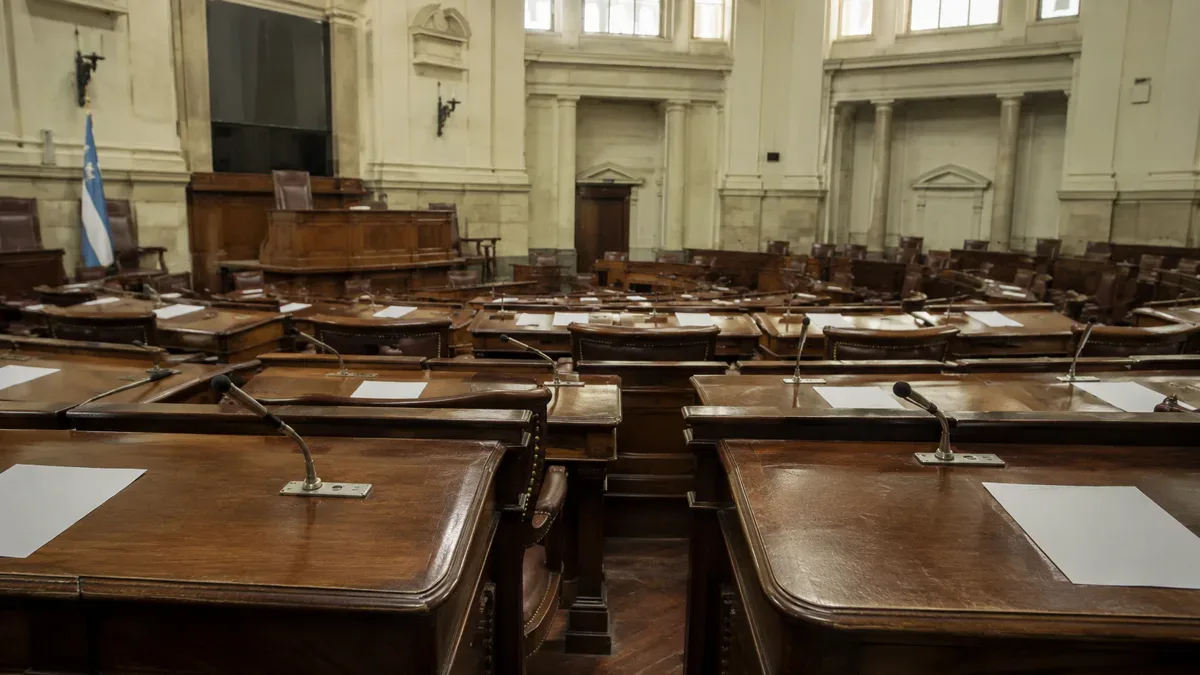 Recinto legislativo vacío con bancas de madera, micrófonos y papeles ordenados sobre los pupitres.
