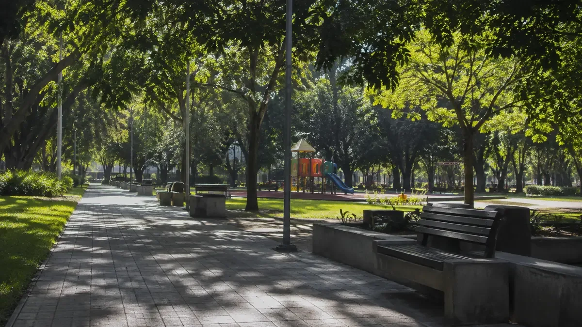 Plaza de barrio con arbolado frondoso, bancos y juegos infantiles vacíos